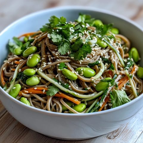 A vibrant Soba Noodle Bowl with crisp cucumber, carrots, and edamame, drizzled with savory sesame dressing on a rustic table.